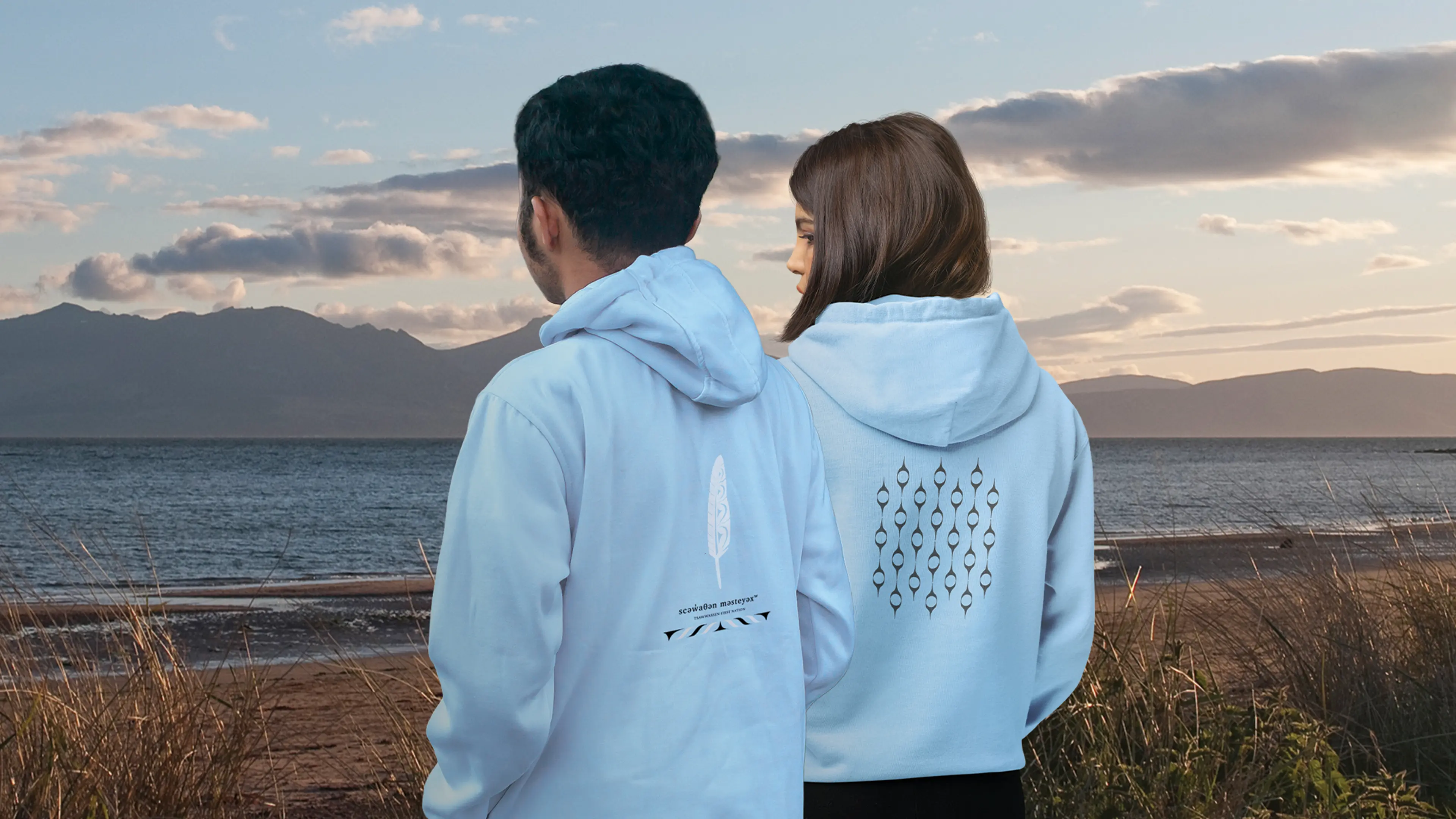 Two people in white hoodies stand on a beach, looking out at the ocean and mountains under a cloudy sky.